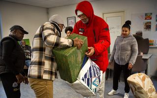 Twee mannen proberen een grote doos in een tas te stoppen. Om hen heen staan verschillende andere mensen toe te kijken.