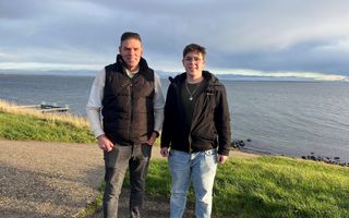 Een man van middelbare leeftijd en een jongen met bril staan op een dijk. Beiden dragen vissersklompen. Achter hen een groot meer.