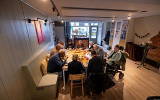 Tien mensen zitten aan een vierkante tafel in een inloophuis te eten.