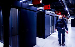 Een man in donkerblauwe bedrijfskleding loopt langs grote computerkasten die achter elkaar in een hal staan opgesteld.