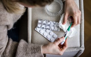 Een vrouw heeft een koortsthermometer in haar handen. Op de leuning van een bank ligt een strip met paracetamol.