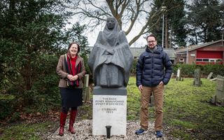 Vrouw met rode trui, bruin jasje en man met blauw jack en bruine broek naast beeld van vrouw op een sokkel.