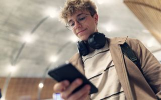 Jongen loopt op overdekt perron met smartphone in zijn hand en koptelefoon om zijn nek.