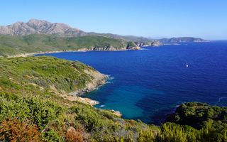 de rotsachtige kustlijn van een eiland in een diepdonkerblauwe zee; de rotsen zijn begroeid met struiken en boompjes; in het water dobbert één zeilboot