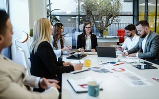 Drie vrouwen en drie mannen zitten om een tafel te vergaderen. Sommigen schrijven, anderen praten.