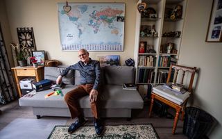 Oudere man met blauw overhemd en bruine broek zit op een grijs bankje. Achter hem aan de muur hangt een wereldkaart. Links van hem staat een boekenkast met allerlei snuisterijen. 