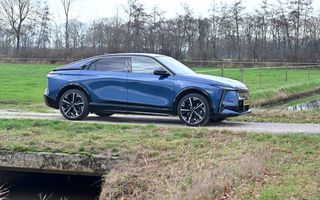 Een blauwe auto staat op een brug in een groen landschap. Op de achtergrond een rij bomen. Net achter de auto paaltjes met wit draad.