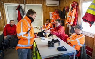 Vijf bouwvakkers rond een witte tafel waarop koffiemokken staan. Drie van de mannen dragen oranje hesjes; een van hen draagt een pet.