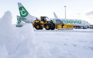 Gele sneeuwschuiver voor een witte landingsbaan waar een wit vliegtuig stilstaat.