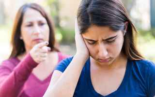 Vrouw met halflang haar en in roze trui steekt haar vinger uit naar haar dochter, een meisje met lang haar en in een blauw shirt. Ze houdt haar rechterhand tegen haar rechteroor en kijkt verdrietig naar beneden. 