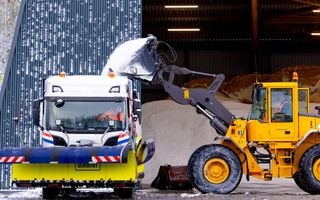 Een gele shovel schept een lading wit strooizout in een witte vrachtwagen met een gele schuiver voorop.