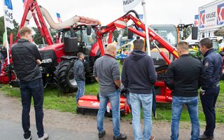 Een groepje mannen staat met de handen in de zakken en de rug naar de camera te kijken naar op een grasveld tentoongestelde rode landbouwmachines.