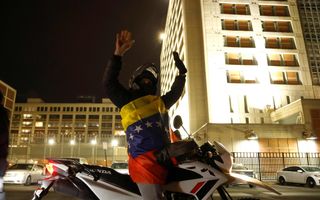 Man gehuld in rood-geel-blauwe vlag zit op motorfiets voor gebouw.