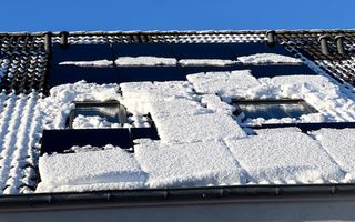 Een laag sneeuw bedekt de zonnepanelen op het schuine dak van een woning, met boven de nok van het dak een strakblauwe lucht.