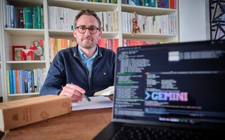 Man aan tafel met laptop en boeken om zich heen. Man draagt bril en heeft een lichte snor en baard.