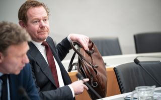 Man in donker pak, wijnrode das, met roodbruin haar en baardje, verplaatst zijn tas van de tafel op de grond. Naast hem zit een ambtenaar die de bewindsman adviseert.