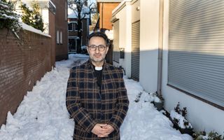 Man met bril, baardje en lange jas staat in de sneeuw naast een gebouw met rolluiken voor de ramen.
