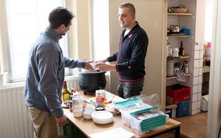 Een man geeft een kom soep aan een andere man. Op de tafel tussen hen in staan kommen, schotels en etenswaren. 