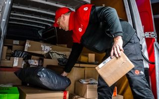 Man in rood-zwarte jas, met rood petje en zwarte broek legt pakketjes in zijn bestelbus. 