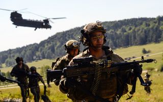 Bewapende mannen in groene camouflagekleding lopen over groene hellingen. Achter hen hangt een chinook in de lucht.