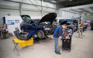 Een bedrijfsruimte met auto’s. Een donkerblauwe auto ligt op een verhoging met de motorkap open. Een man in een blauw jack staat bij een zwarte kist op wielen. 