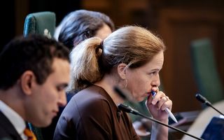 Een vrouw met het haar in een staart zit met een pen voor haar mond achter een microfoon. Naast haar zit een man.