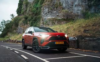 Rode, hoge auto staat met koplampen aan op een parkeerplaats naast een asfaltweg. 