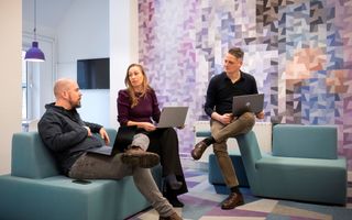 Twee mannen en een vrouw met laptops op schoot, zittend op bankjes. Ze zijn in overleg met elkaar. 