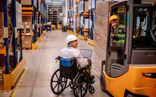 Een man in een rolstoel overlegt met een vrouwelijke collega die in een heftruck zit, in een grote bedrijfshal.