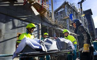 Mensen in gele hesjes en met gele helmen op vervoeren een brancard. Daarop ligt iemand onder een lichtblauw zeil.