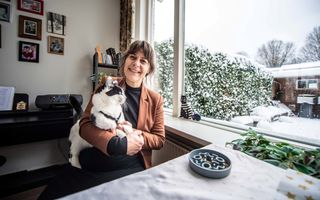 Vrouw zit met kat op schoot voor raam van haar woonkamer.