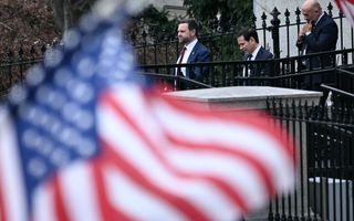 Drie mannen in pak lopen langs een donkergekleurd hek. Op de voorgrond is een Amerikaanse vlag zichtbaar. 