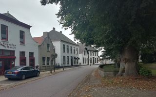 Foto van een rij witte panden langs een klinkerweg met een flauwe bocht. Aan de overkant van de weg staan dikke bomen.