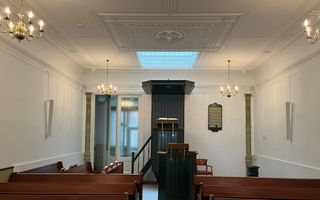 Klein kerkinterieur met witte wanden en bruine, houten banken. Centraal tegen de achterwand een donkergeverfde kansel.