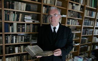 Predikant met wit haar en bril in zwart pak met stropdas staat voor boekenkast.