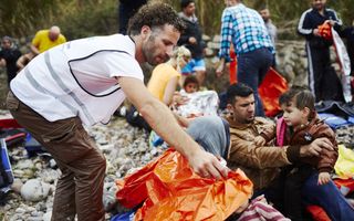 Een man met een wit hesje en een bruine broek slaat een oranje plastic zeiltje om de schouders van een vrouw met een kind in haar armen. Om hen heen zitten andere mensen.