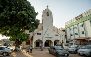 wit kerkgebouw op NIgeriaanse zandgrond, op de voorgrond oude auto’s en mensen in gekleurde kleding die richting de kerk lopen