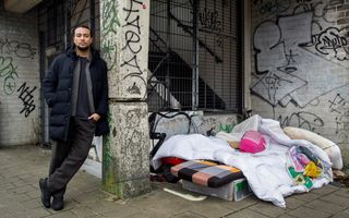 Getinte man leunt tegen een betonnen paal waarop graffiti is gespoten. Ernaast ligt op de straat een matras met dekbed en kussens.
