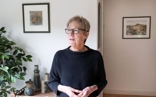 Vrouw met kort grijs haar en bril, in een huiskamer waarin kleine schilderijen hangen.
