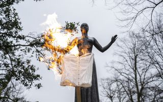 Vlammen slaan uit een korset, dat met een stok omhoog wordt gehouden. Op de achtergrond een standbeeld van een vrouw.