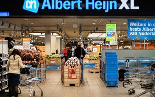 Ingang van een supermarkt van Albert Heijn. Een vrouw loopt achter een winkelwagen. Er zijn blauwe mandjes te zien en, verder in de winkel, mensen, bloemen en groenten.
