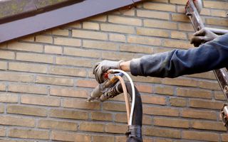 Twee slangen met spuitmond gaan in een bakstenen muur, terwijl een hand de slangen vasthoudt.