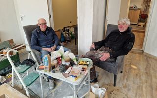 Twee mannen in klusoutfit zitten achter een tafeltje dat vol staat met etenswaren. In de ruimte waarin ze zitten staan allerlei klusattributen.