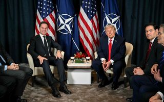 Twee mannen in pak zitten op een stoel voor een rijtje vlaggen, van de VS en van de NAVO. 