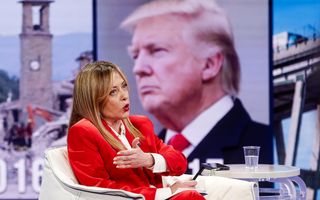 Een vrouw in een rood mantelpakje spreekt. Op de achtergrond is een afbeelding van een man met blond haar zichtbaar.