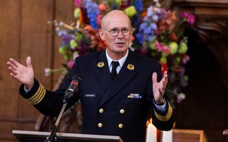 Man in donkerblauw marine-uniform achter katheder. Breidt, al sprekend, zijn handen uit. 