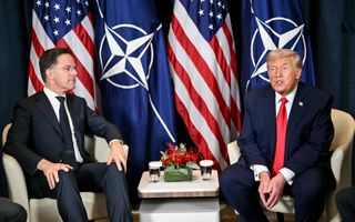 Twee mannen in donkere maatpakken zitten ieder op een crèmekleurige stoel, met achter hen verschillende vlaggen.