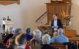 Man met een bril, jasje, spreekt de mensen in de kerkbanken vanaf het podium toe.