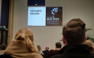 ”Van harte welkom, bid mee” staat op de muur geprojecteerd. De mensen die er zitten zie je op de rug.