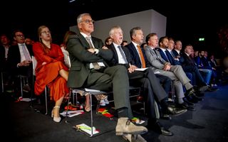 Mensen, meest mannen in pak, zitten in rijen in een zaal tegen een donkere achtergrond. Op de tweede rij zit een vrouw in felrode jurk.